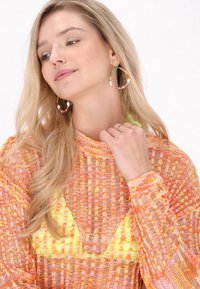 Maglione a rete colorato con tonalità di arancione, rosa e giallo, abbinato a grandi orecchini a cerchio con perline. Il modello fa un gesto con la mano.