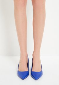 Dei tacchi alti in pelle blu con design a punta. Texture liscia e stile minimalista, presentati su un paio di gambe nude.