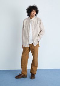 Lys beige stribet skjorte med knapper lagdelt over en hvid t-shirt, parret med brune vide bukser og mørkebrune sko med frynser.