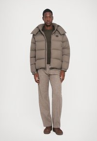 Male wearing taupe puffer jacket, brown knit sweater, gray wide-leg trousers, and brown sneakers standing against plain background.