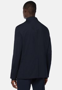 Marinblå skräddarsydd kappa med hög krage, strukturerade axlar och slät yta. Inkluderar slits bak och minimalistiska sömmar för ett rent utseende.