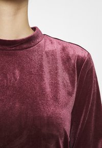 Burgundské sametové tričko s vysokým výstřihem, hladkou strukturou a jemným leskem. Disponuje dlouhými rukávy a těsným střihem.