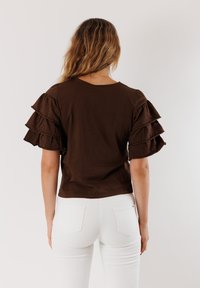 Top in cotone marrone con maniche a strati e volants. Presenta una scollatura rotonda, maniche corte e un design aderente. Indossato con jeans bianchi.