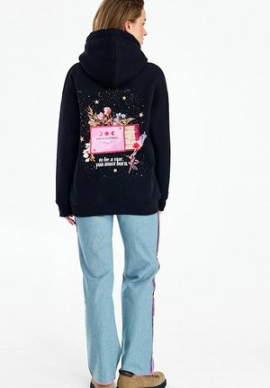 Kobieta w czarnej bluzie z kapturem z nadrukiem kwiatów, gwiazd i tekstem "Pozostań w swojej magii, aby być gwiazdą, musisz płonąć", w zestawieniu z jasnoniebieskimi spodniami.