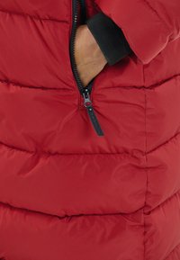 Röd puffjacka med kviltade sektioner. Har en dragkedjeficka och svart dragkedjeskäle. Tyget verkar slätt med en lätt glänsande yta.