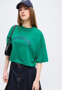 Νεαρή γυναίκα με πράσινο κοντό T-shirt Calvin Klein, σκουρόχρωμη τζιν φούστα, μπλε καπέλο και μαύρη τσάντα ώμου σε απλό φόντο.