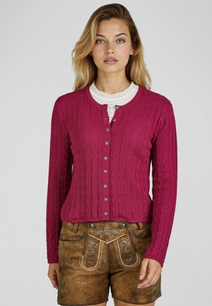 Junge Frau trägt eine knopfgebundene magentafarbene Strickjacke mit Zopfmuster über einem weißen Spitzenoberteil und braunen, bestickten Lederhosen.