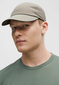 Jasnoszara czapka o teksturowanej powierzchni, z zaokrąglonym daszkiem i detalem z logo; noszona z zieloną koszulą z krótkim rękawem, prezentującą minimalistyczny design.