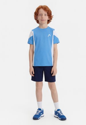 Punase juustega poiss seisab näoga ettepoole, kandes sinise ja valge spordisärgi, tumerohelisi lühkse, valgeid sokke ja siniseid tossusid valgel taustal.