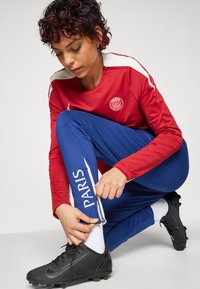 T-shirt de sport rouge avec des accents blancs, logo sur la poitrine. Pantalon bleu avec le texte "PARIS", chaussures de football noires. Fond gris.