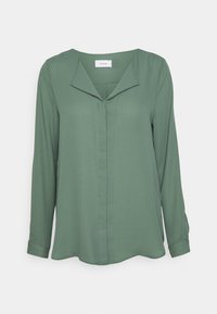 Blouse verte à manches longues avec un décolleté en V, présentant une coupe décontractée, une texture lisse et un léger brillant. Des coutures notables sont visibles sur le devant.