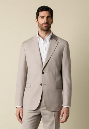 Man met donker haar, gekleed in een lichtbeige pak en een wit overhemd, staand tegen een effen beige achtergrond.