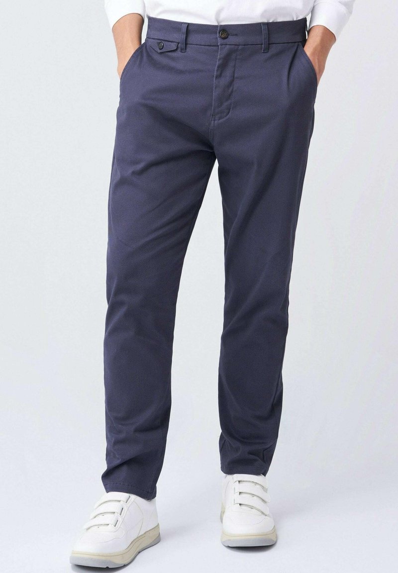 Homme portant un pantalon slim bleu marine, les mains dans les poches et des baskets blanches, debout devant un fond clair uni.