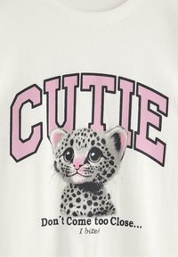 Hvid bomulds t-shirt med en tegneserieleopard med store øjne, sorte pletter og en lyserød næse. Teksten "CUTIE" står i fede lyserøde bogstaver ovenfor.