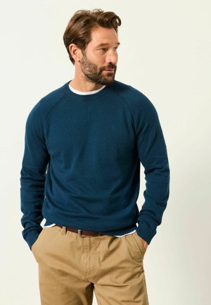 Mann mit braunen Haaren und Bart, der einen blauen Rundhalspullover und beige Hosen trägt, die Hände in den Taschen, schaut nach rechts.