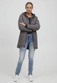 Grå polstret jakke med hette, glidelås og frontlommer. Brukt over en svart skjorte og kombinert med lyseblå jeans og hvite sneakers.