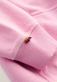 Felpa con cappuccio rosa chiaro con polsino a coste e etichetta logo arancione e blu navy. Tessuto morbido, trama liscia e design con tasca a marsupio.