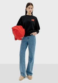 Jonge vrouw in een zwart sweatshirt met een rood berlogo, blauwe jeans, beige slippers, met een rode tas tegen een effen achtergrond.