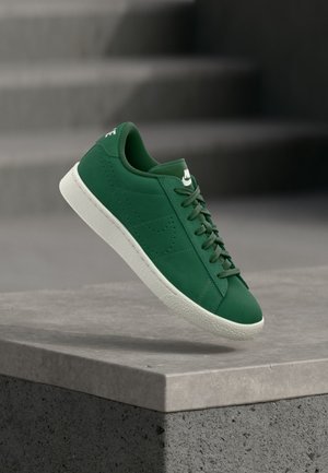 Baskets Nike vertes avec semelle et lacets blancs, flottant au-dessus d'un rebord en béton avec des marches floues en arrière-plan.