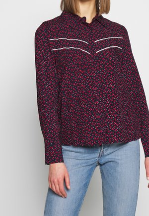 Frau trägt ein langärmeliges marineblaues Shirt mit rotem Blumenmuster, weißen horizontalen Nähten auf der Brust, kombiniert mit hellblauen Jeans.