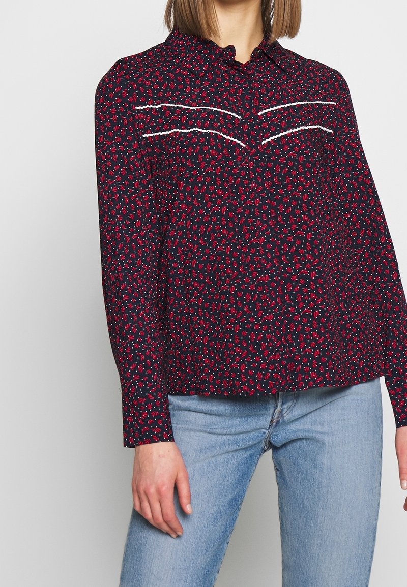 Femme portant un t-shirt bleu marine à manches longues avec imprimé floral rouge, surpiqûres blanches horizontales sur la poitrine, associé à un jean bleu clair.