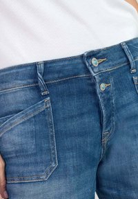 Jeans en denim bleu avec une taille mi-haute, fermeture à double bouton, détails de poches avant et couture contrastée. Texture douce.