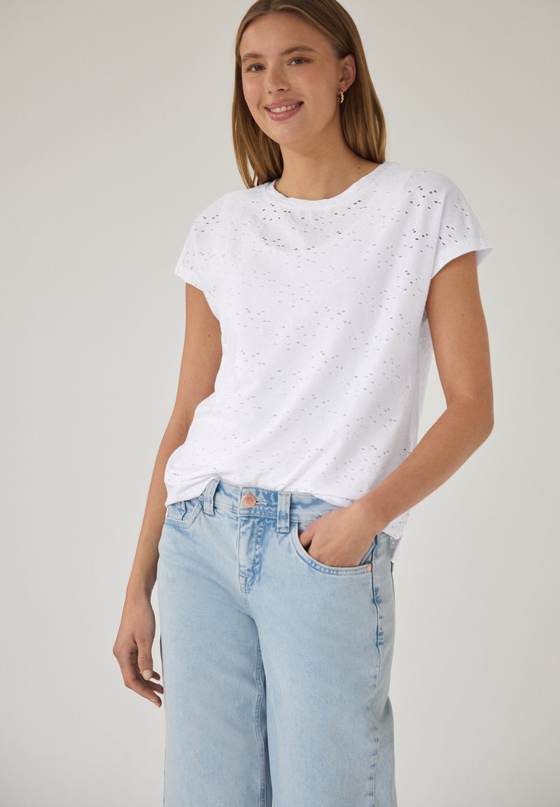 Joven mujer con una camiseta blanca perforada de manga corta y vaqueros azul claro, sonriendo con una mano en el bolsillo.