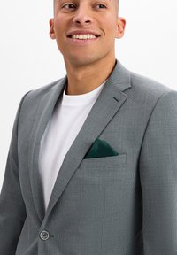 Szary w kratę marynarka z wykładanymi klapami, jednolitym zapięciem na guzik oraz poszetką w ciemnozielonym kolorze. Noszona na białej koszulce.