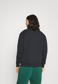 Hanorac negru cu manșete și tiv cu nervuri, potrivire standard, purtat deasupra pantalonilor verzi. Prezintă un decolteu rotund și un design minimal.
