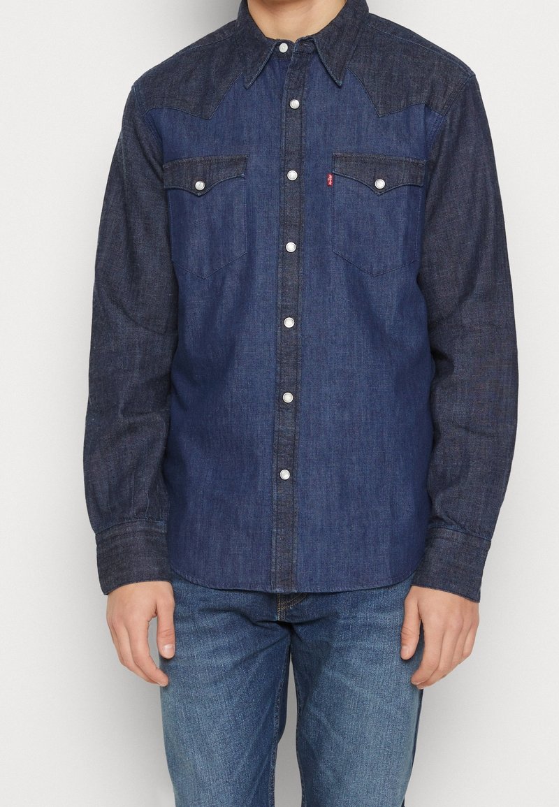 Camisa de denim con tonos contrastantes, cierre de botones en la parte delantera, dos bolsillos en el pecho, diseño de yugo puntiagudo y acentos de hardware plateado.