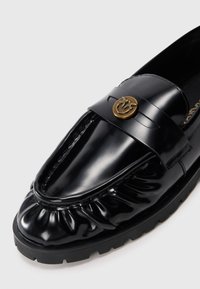Mustade patendi nahast loaferid kogunenud varbaga detailiga, paksu kummitald ja kuldne ümmargune emblem laial varvaste rihma peal.