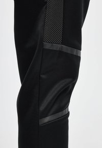 Pantalones deportivos negros con un acabado liso, que cuentan con un panel de acento texturizado y rayas negras mate a lo largo de las costuras laterales.