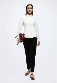 Άσπρο εφαρμοστό πουκάμισο, μαύρο στενό παντελόνι και μπορντό τσάντα. Το μοντέλο φορά μπορντό μπαλαρίνες, στέκεται μπροστά σε απλό λευκό φόντο.