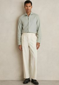 Cămașă verde deschis, cu nasturi, cu un finisaj texturat, purtată cu pantaloni albi plisați și pantofi negri. Design simplu cu culori delicate.