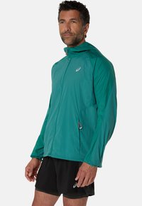 Veste légère teal avec capuche, dotée d'une fermeture éclair à l'avant, de poches latérales et d'un logo réfléchissant. Texture lisse et coupe athlétique.