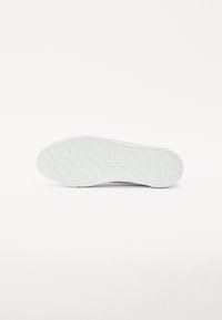 Hvit gummisåle med vertikale riller for bedre grep, med en rund tå og logo detalj i midten. Rent, minimalistisk design.