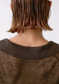 Brun sweater med en bred, mørk ribbestrikket halslinie, fremstillet af et tekstureret, let gennemsigtigt stof og en afslappet pasform. Fugtigt, skulderlangt hår er synligt.