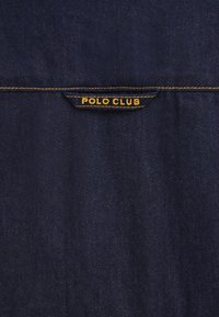 Mørkeblåt denimstof med subtil tekstur. Har et rektangulært mærke syet øverst med teksten "POLO CLUB" i gult.