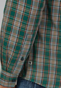 Camisa de cuadros verde con una mezcla de líneas naranja, beige y azul, puños con botón y una pequeña etiqueta cosida cerca del dobladillo.