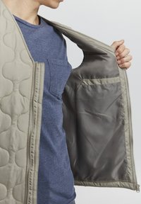 Padded beige vest with quilted exterior, shown unzipped to reveal a smooth gray interior and a small pocket on the inner lining.