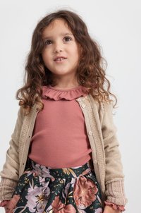 Cardigan beige lavorato a maglia con dettagli intricati, indossato su una blusa rosa con ruches, abbinato a una gonna floreale nei toni del verde scuro, rosa e con accenti dorati.