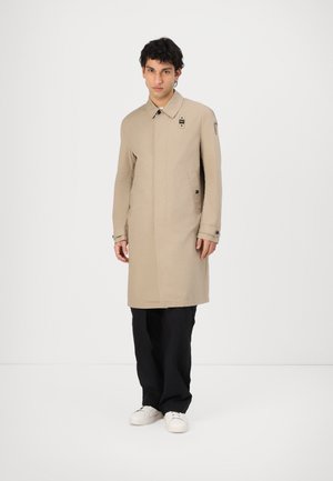 Beige lang frakke lavet af letvægtsstof, med høj krave, sidelommer og knapdetaljer. Sammen med sorte bukser og hvide sko.