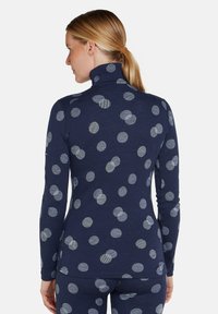 Frau in einem marineblauen Langarmoberteil und Hosen mit großen hellblauen Punkten, von hinten vor einem einfarbigen Hintergrund gezeigt.