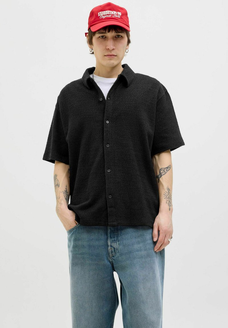 Sort kortærmet skjorte med knapper i struktureret stof, parret med lyseblå jeans. Modellen har en rød kasket på med hvid tekst.