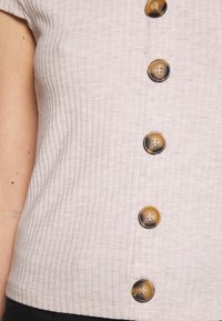 Lys beige ribbet kortærmet bluse med en central knapplet med store runde brune knapper, glat tekstur og lige kant.