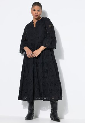 Schwarzes Maxikleid mit Augenstickerei, mit schichtigem Design und Glockenärmeln. Getragen mit schwarzen, mit Nieten besetzten, Knöchelstiefeln, die einen strukturierten Look zeigen.