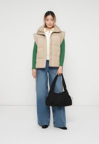 Ženska oblečena v bež puhovko čez zelene dolge rokave in belo majico, široke modre jeans hlače, drži črno torbico na enobarvni beli podlagi.