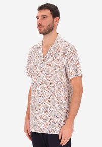 Uomo che indossa una camicia a maniche corte con un motivo floreale multicolore e pantaloni scuri, in piedi contro uno sfondo bianco semplice.