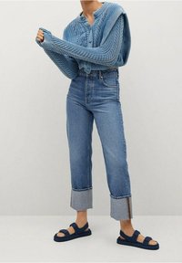 Pull en tricot bleu avec un motif texturé, jean en denim clair taille haute avec revers, et sandales bleu marine sur pieds nus.