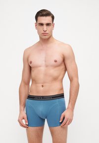 Un uomo che indossa boxer in cotone blu con un elastico nero che presenta il nome del marchio "JACK & JONES", in piedi davanti a uno sfondo semplice.